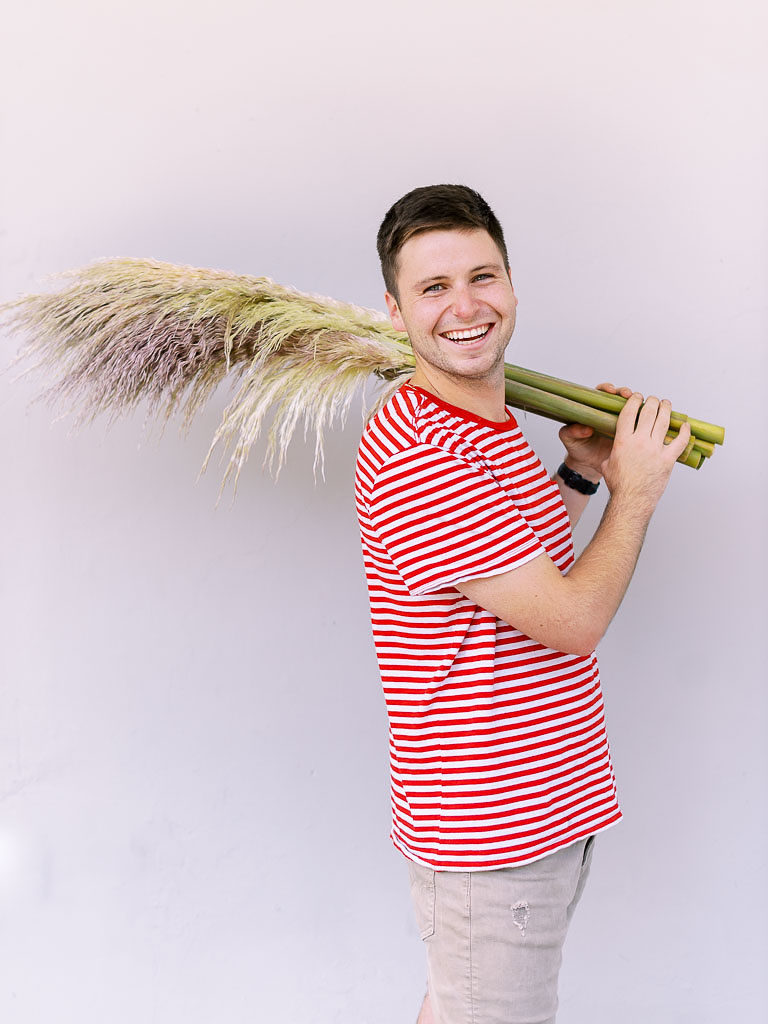 Young man in striped red shirt laughing and holding fan-like plant over his shoulder at floral workshop in Charleston, SC