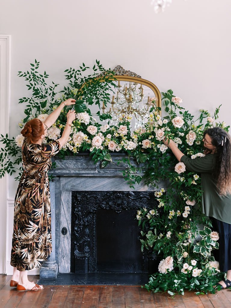 Two women arranging a floral installation over a gray fireplace mantle and large gold-edged mirror by floral workshop photographer Kim Branagan