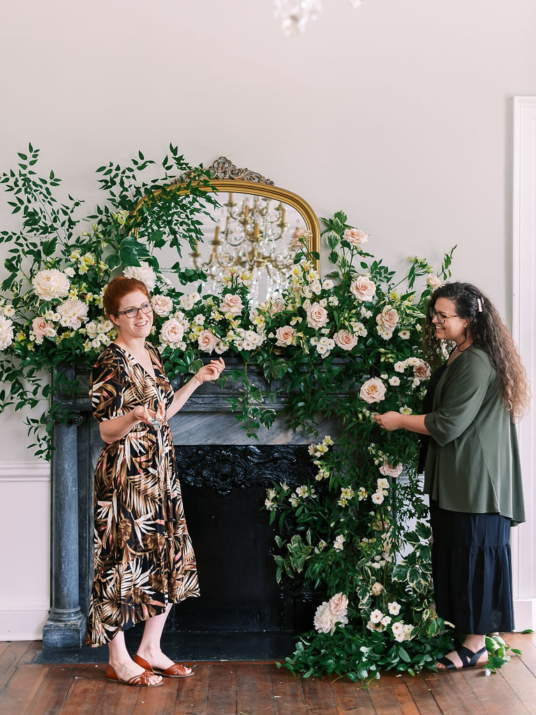 Two women talking and smiling while they create floral display of carnations and greenery over a gray fireplace and gold-edged mirror