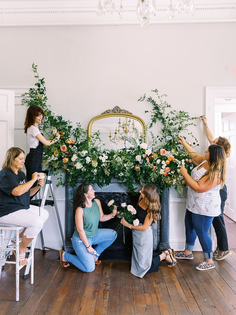 Six women working on an installation of greenery and carnations above a fireplace by floral workshop photographer Kim Branagan