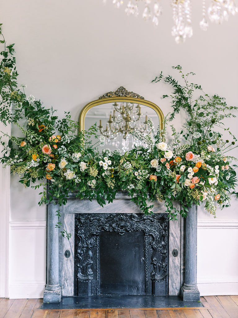 A completed floral workshop installation of greenery and carnations around a gold-edged mirror on top of a silver and black fireplace
