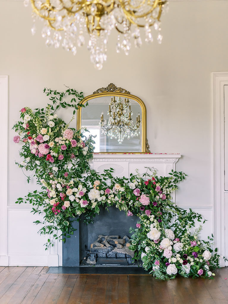Finished floral installation on a fireplace with a chandelier in the foreground that is reflected in the gold-edged mirror on top of the fireplace
