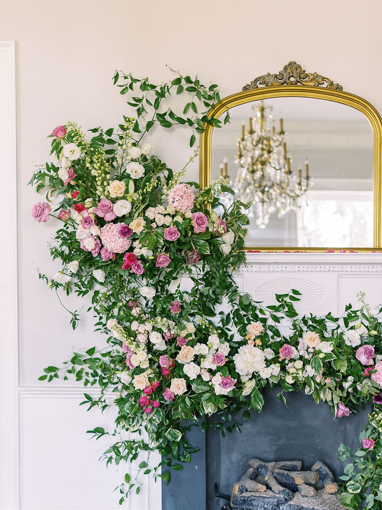 An arrangement of flowers and greenery on a fireplace beside a gold-edged mirror reflecting a chandelier photographed by floral workshop photographer Kim Branagan