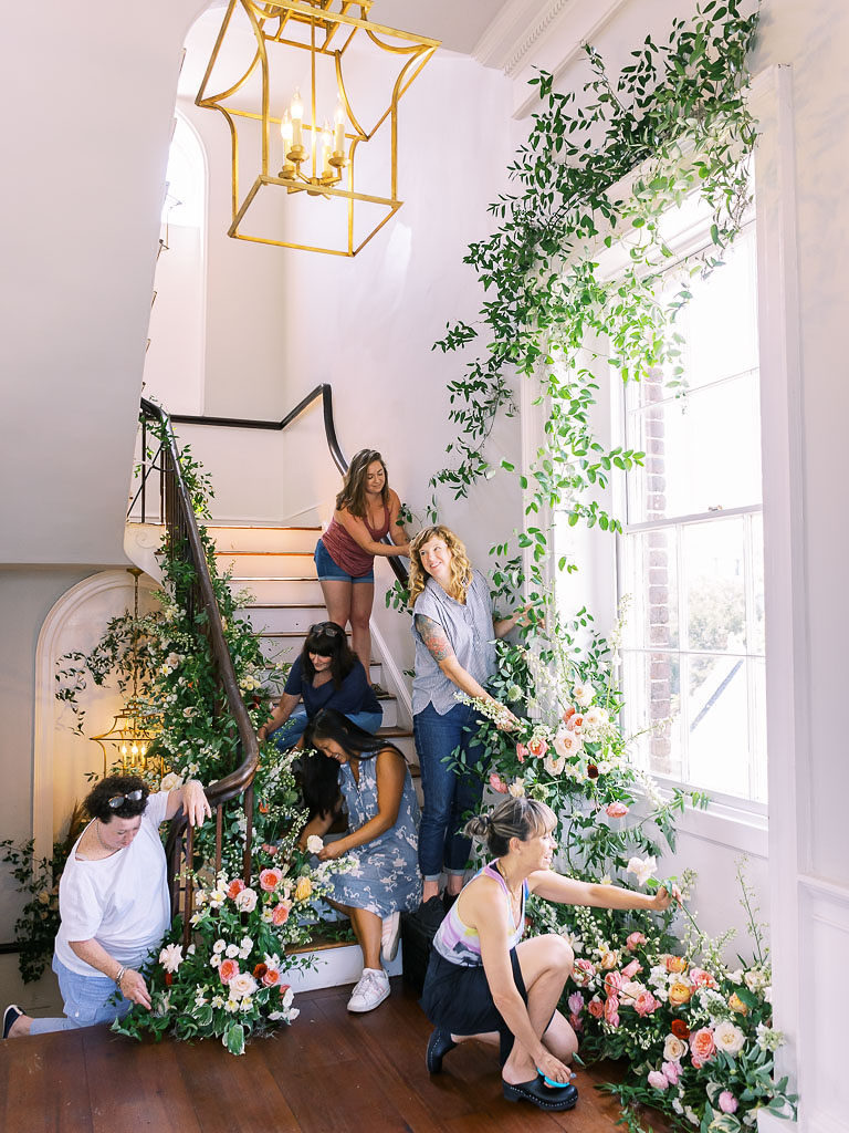 Six women working on a floral installation surrounding a large white-framed window and a curving bannister for a short flight of white stairs