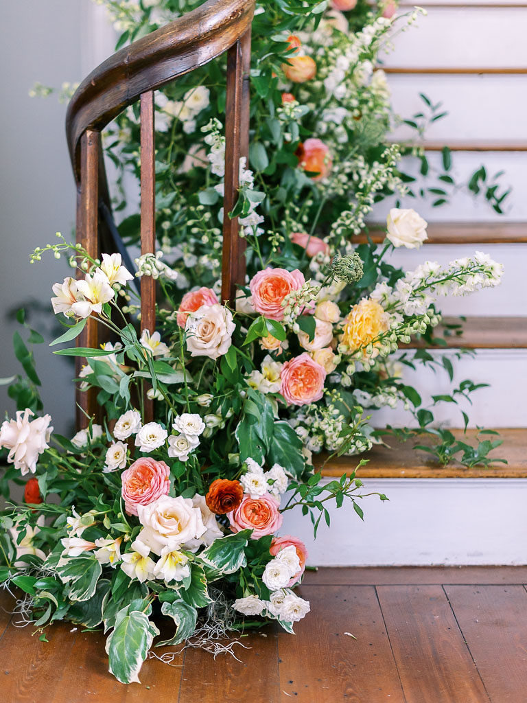Close-up of yellow, peach, and orange flowers and greenery arranged around a dark brown curving bannister by some wooden stairs photographed at a floral workshop