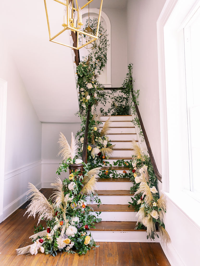 Finished installation of greenery, tan fan-like plants, and flowers along a flight of wooden stairs by floral photographer Kim Branagan