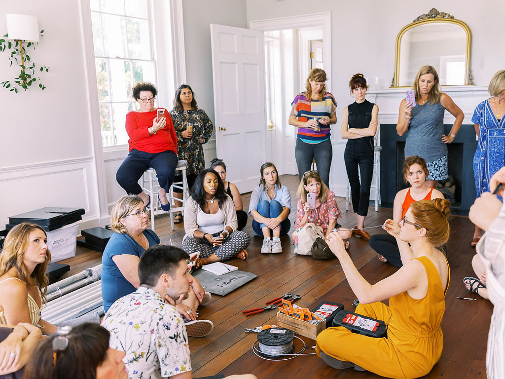 A group of people standing and sitting on a wooden floor while watching a woman in a mustard yellow jumper demonstrate how to do a floral installation