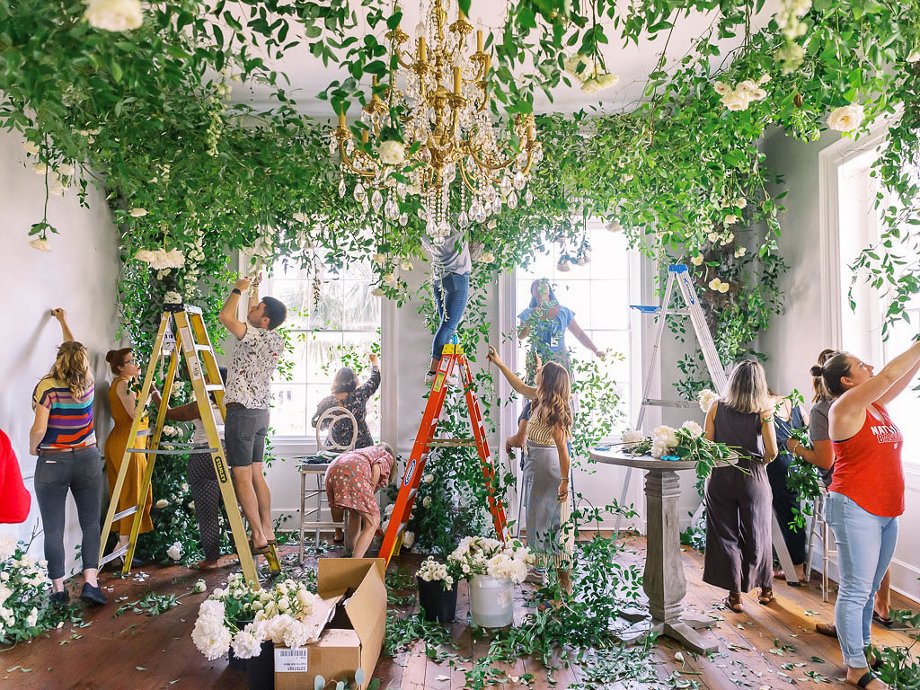 Floral designers working on arranging greenery by the ceiling and around several large windows in a room with a chandelier and wooden floor at a floral workshop