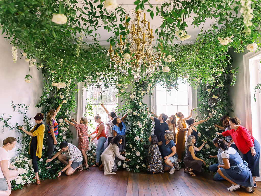Floral designers arranging flowers in greenery in installations surrounding a chandelier and several large windows photographed by floral photographer Kim Branagan