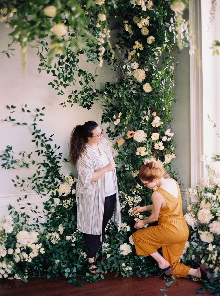 Two women arranging greenery and light yellow flowers along a white wall by a window at The Foraged Collective Workshop in Charleston