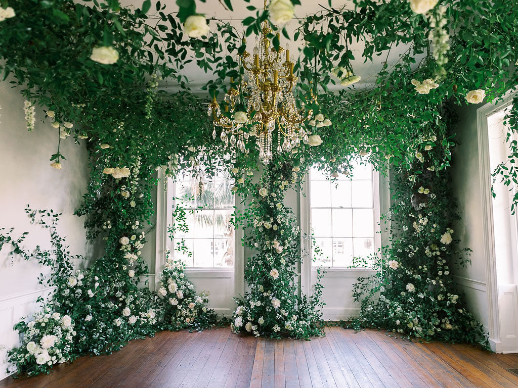 A finished floral installation of greenery and white and light yellow flowers surrounding two tall windows and a chandelier by floral photographer Kim Branagan