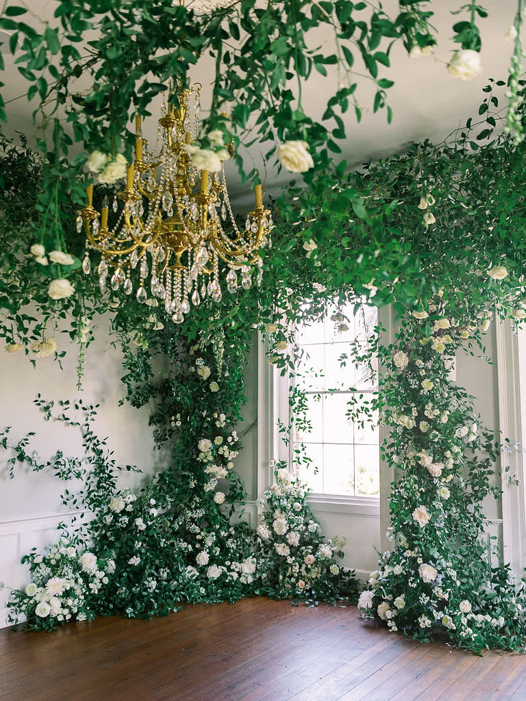 An angled view of an installation at a floral workshop involving greenery and flowers around a chandelier and a large window