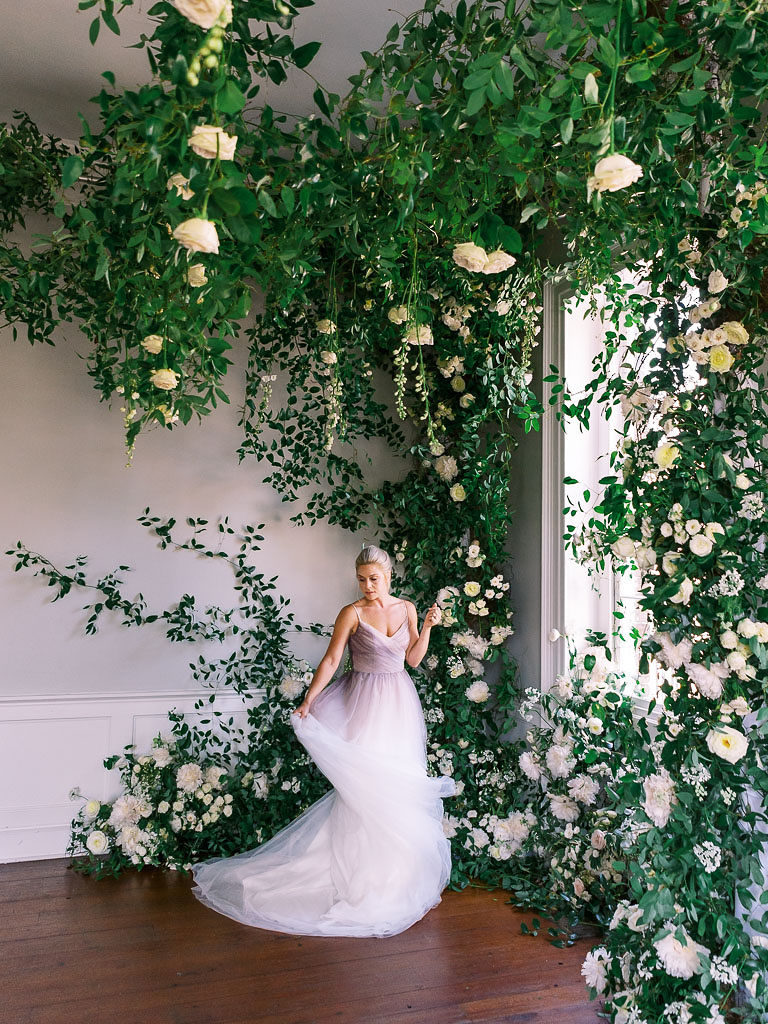 Woman in lavender gown standing by a floral installation of pastel flowers and greenery around a large window by wedding photographer Kim Branagan