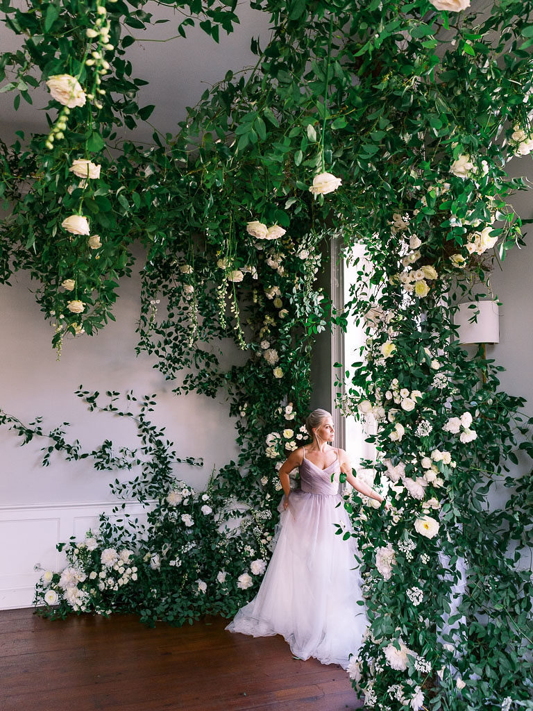 Distant shot of woman looking out a large window and surrounded by greenery and flowers