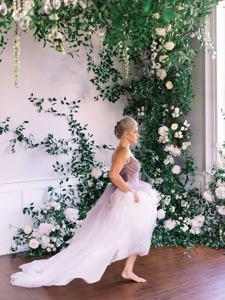 Woman holding up train of lavender gown and walking barefoot toward window in front of floral installation against a white wall