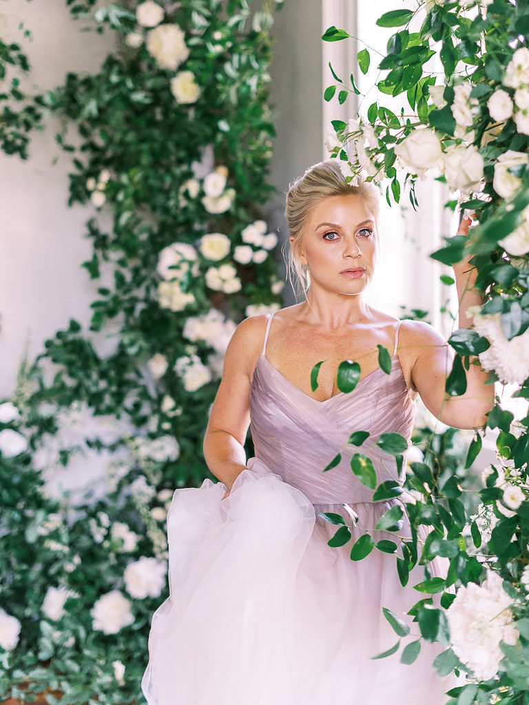 Blonde woman in lavendar gown standing by window surrrounded by white and light yellow flowers and greenery
