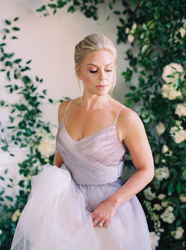 Blonde woman holding train of lavender gown, looking down, and standing in front of floral installation photographed by wedding photographer Kim Branagan