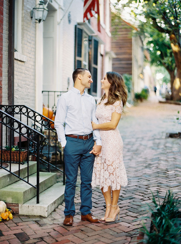 A woman holds on to her fiance's arm while they gaze into each other's eyes and smile on a brick sidewalk in a neighborhood in Old Town Alexandria, Virginia. Photographed by DC wedding photographer Kim Branagan.