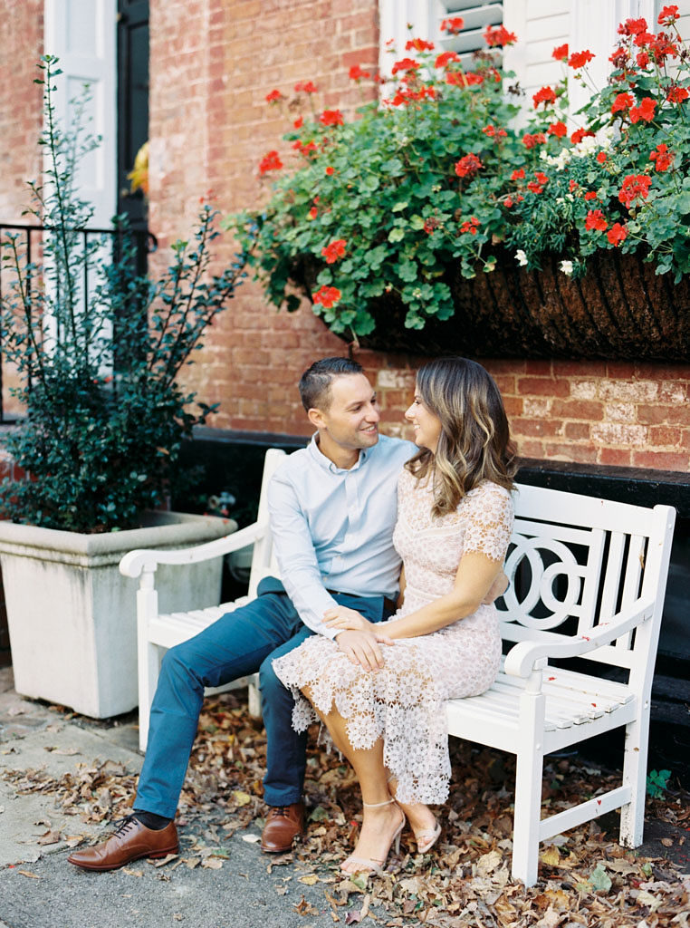 An engaged couple sits close to each other, gazing into each other's eyes. They are sitting on a white wooden bench in front of a brown brick building with red flowers in the window sill in Old Town Alexandria, Virginia. Photographed by Alexandria, Virginia commercial photographer Kim Branagan.
