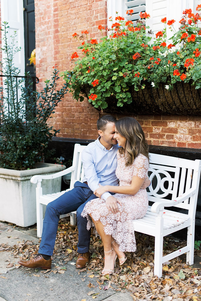 An engaged couple sits close to each other, laughing, on a white wooden bench in front of a red brick building with red flowers in the window sill in Old Town Alexandria, Virginia.