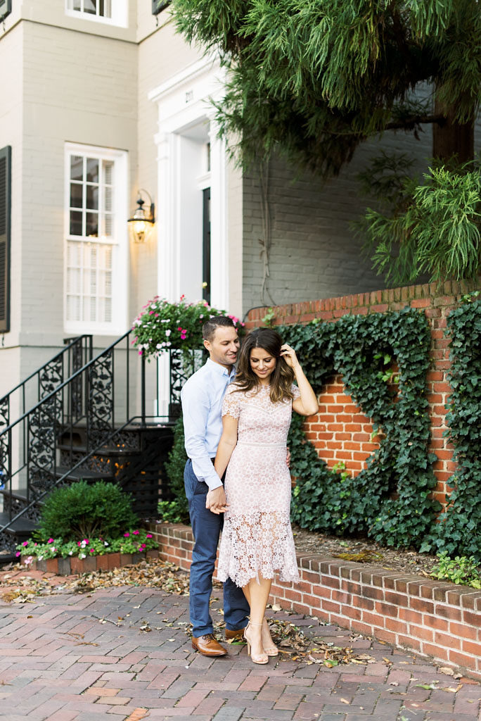 An engaged couple holding hands and smiling in front of a Victorian home with a brown brick walkway, brick wall covered in greenery and trees in the background in Old Town Alexandria, Virginia.