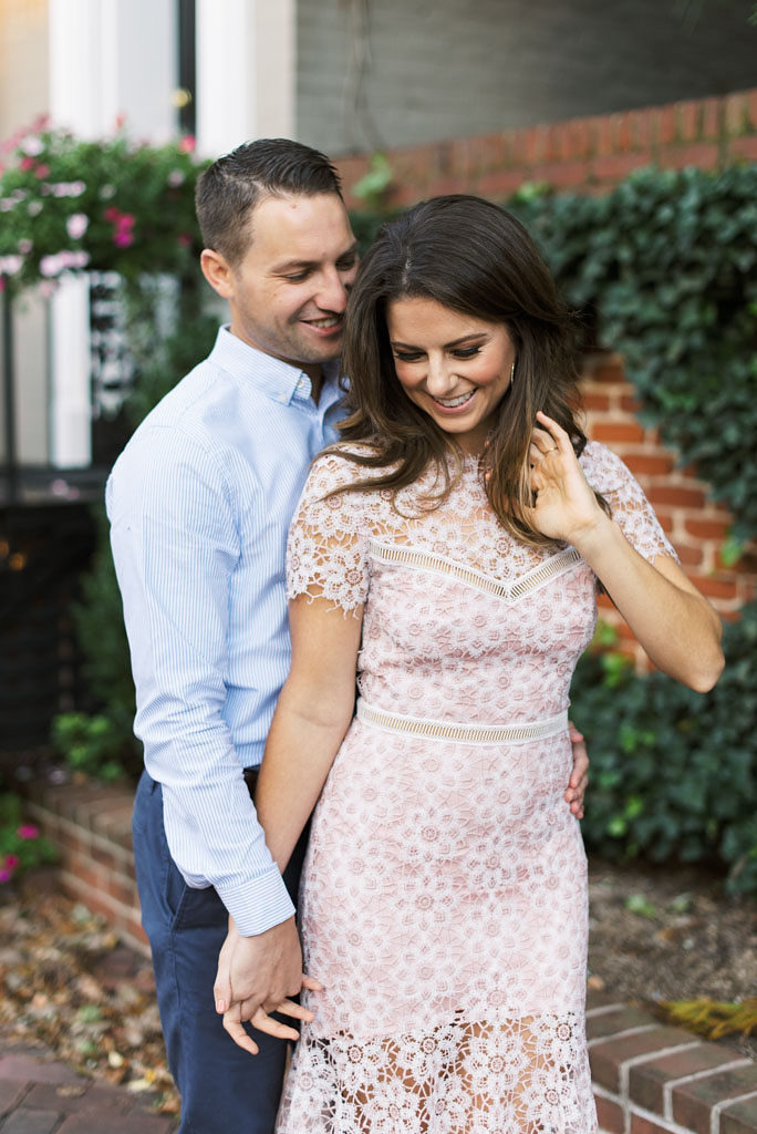 A man stands behind his fiance, leaning toward her, smiling, and holding her hand. His fiance stands in front of him, with her hand in her hair and wearing a light pink dress. They are standing in front of a brown brick wall with shrubbery and fallen leaves on the ground. Photographed by Virginia wedding photographer Kim Branagan.