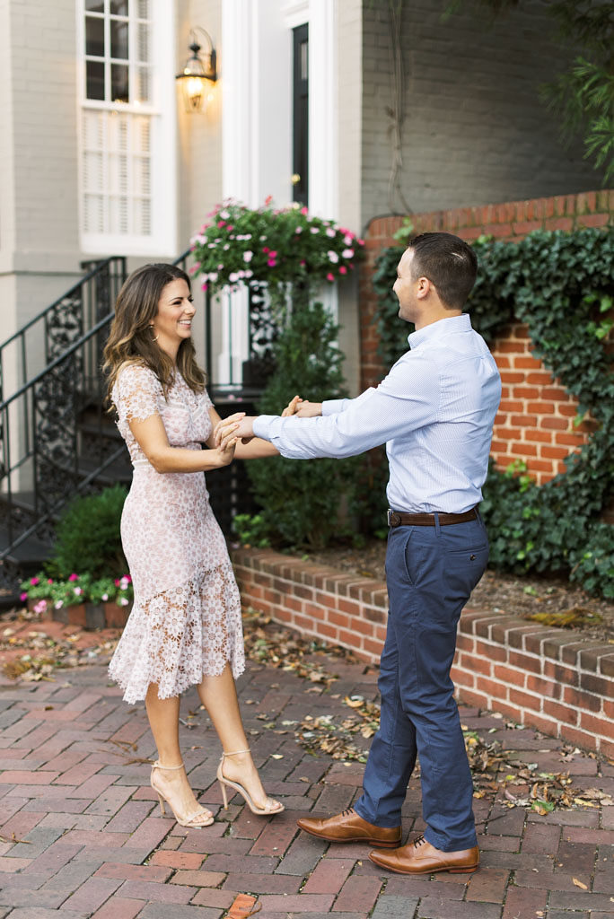 An engaged couple stands facing each other about one foot apart. They are holding hands and smiling in front of a historic gray home, a brick wall with ivy and flowers on it, and brick walkway in Old Town Alexandria, Virginia. Photographed by DC wedding photographer Kim Branagan.