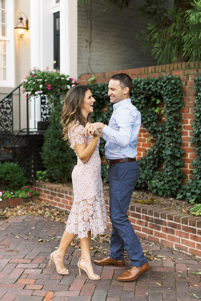 An engaged couple dances, holding hands and laughing, by a historic gray building with white trim, a brown brick wall, and brick walkway in Old Town Alexandria, Virginia. Photographed by DC commercial photographer Kim Branagan.