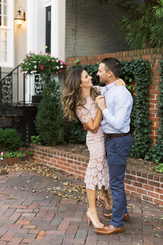 An engaged couple slow dancing and smiling in front of a gray building with white trim, a brown brick wall, and brick walkway. Photographed by Virginia wedding photographer Kim Branagan.