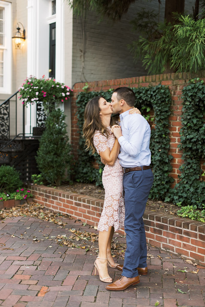 An engaged couple stands facing each other in front of a gray building with white trim and brown brick wall and kisses. They are holding hands.