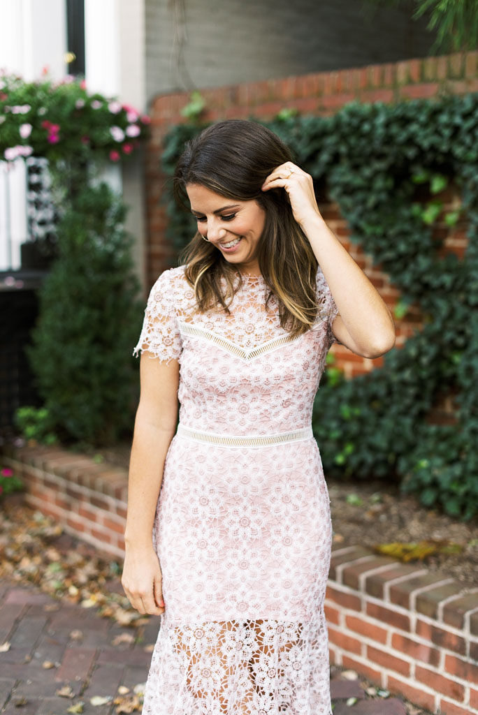A smiling woman wearing a pink lace dress brushes her hand through hair and looks toward the ground. She is standing in front of an ivy-covered brick wall in Old Town Alexandria, Virginia. Photographed by DC wedding photographer Kim Branagan.