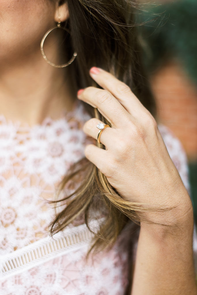 A closeup photo of a woman with her hand in her hair, with a diamond engagement ring on her hand. She has long golden brown hair and is wearing a pink lace top.