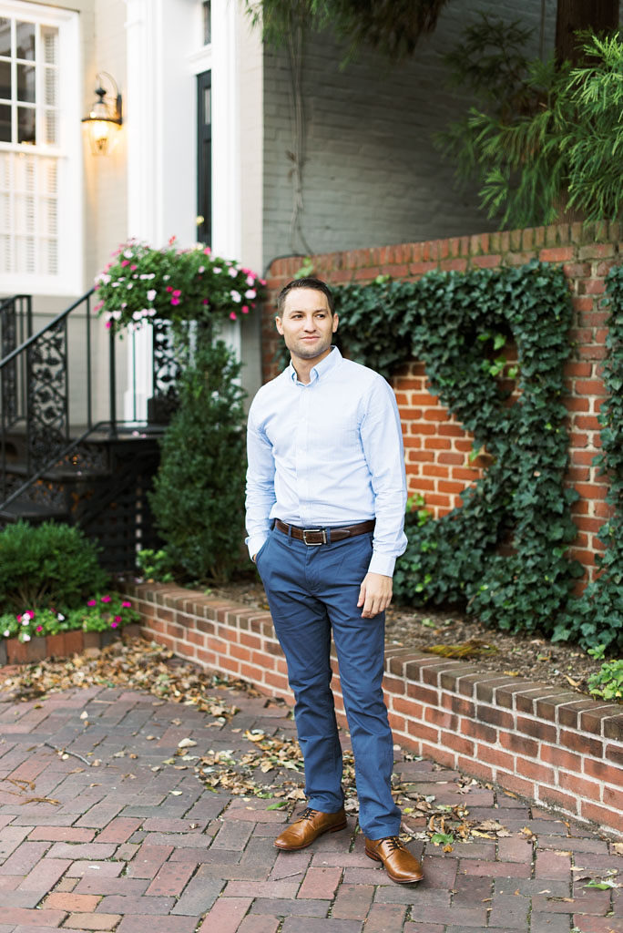 A man in a light blue shirt, navy blue slacks, and brown dress shoes stands on a brick walkway in front of a brown brick wall with ivy. He is looking off to his side.