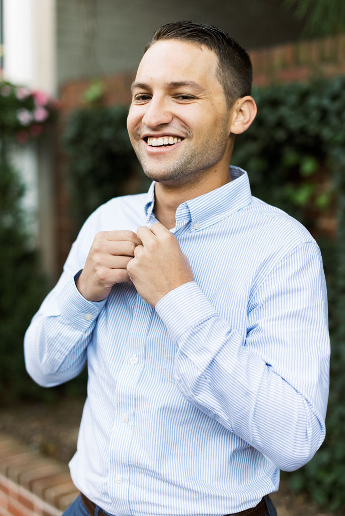 A man wearing a white button down shirt with light blue stripes buttons the top button of his shirt and smiles while standing in front of green shrubbery in Old Town Alexandria, Virginia. Photographed by Alexandria, Virginia Photographer Kim Branagan.