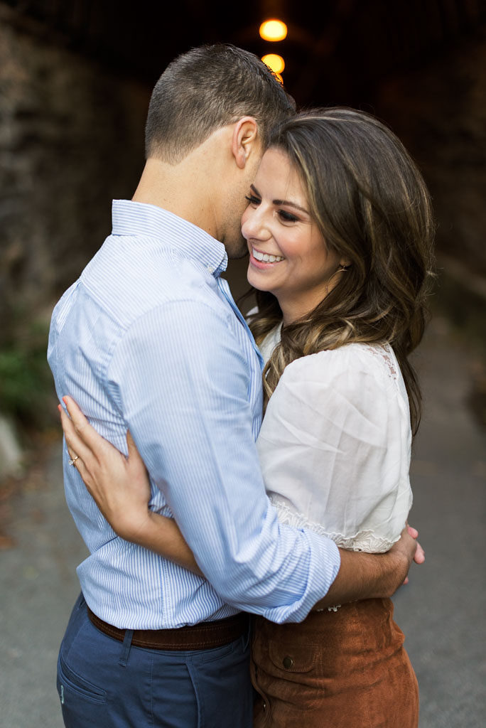 A young engaged couple hugs each other in front of a dark background. The man is looking away from the camera, and the woman is smiling.