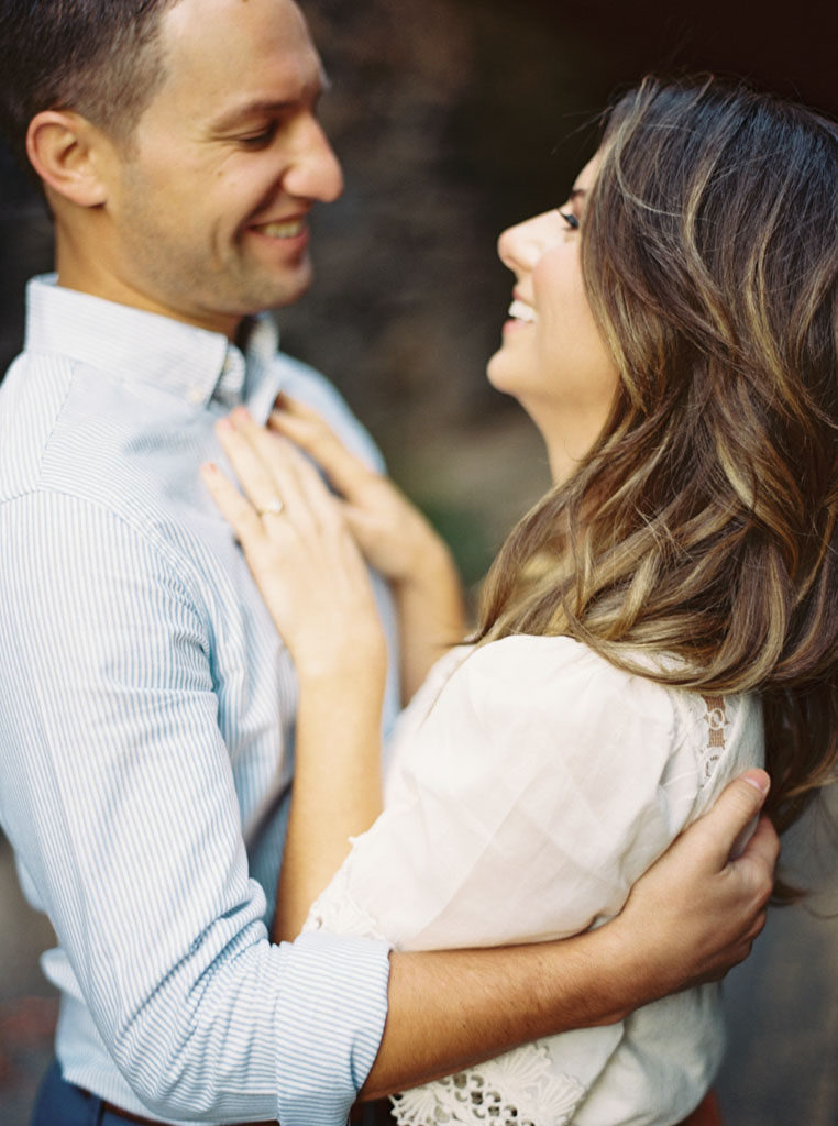 A man in a striped, light blue button down shirt holds his fiance. Her hands are on his chest and she is looking up at him, smiling. Photographed by Alexandria, Virginia wedding photographer Kim Branagan.