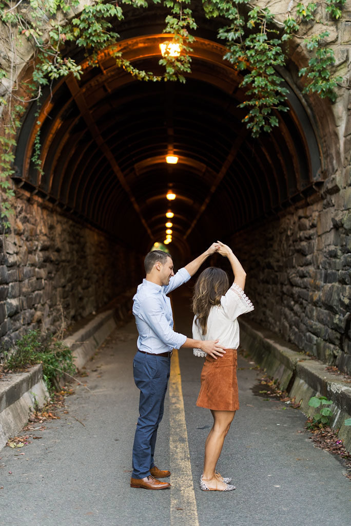 A man twirling his fiance while dancing in front of a historic brick tunnel with lights in Old Town Alexandria, VA.