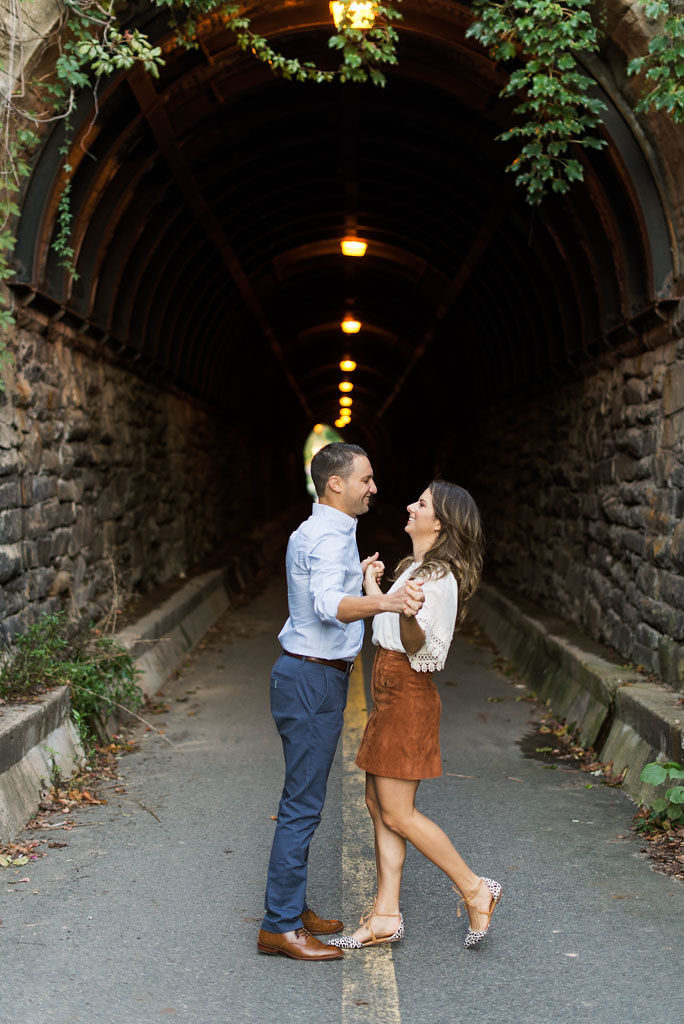An engaged couple standing in front of a brick tunnel with green foliage in Old Town Alexandria, Virginia, holding hands and laughing. Photographed by Virginia wedding photographer, Kim Branagan.