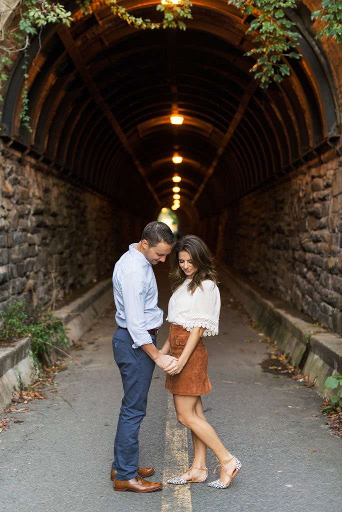 An engaged couple facing each other and holding hands stands in front of a brick tunnel with green vines hanging above the tunnel entrance