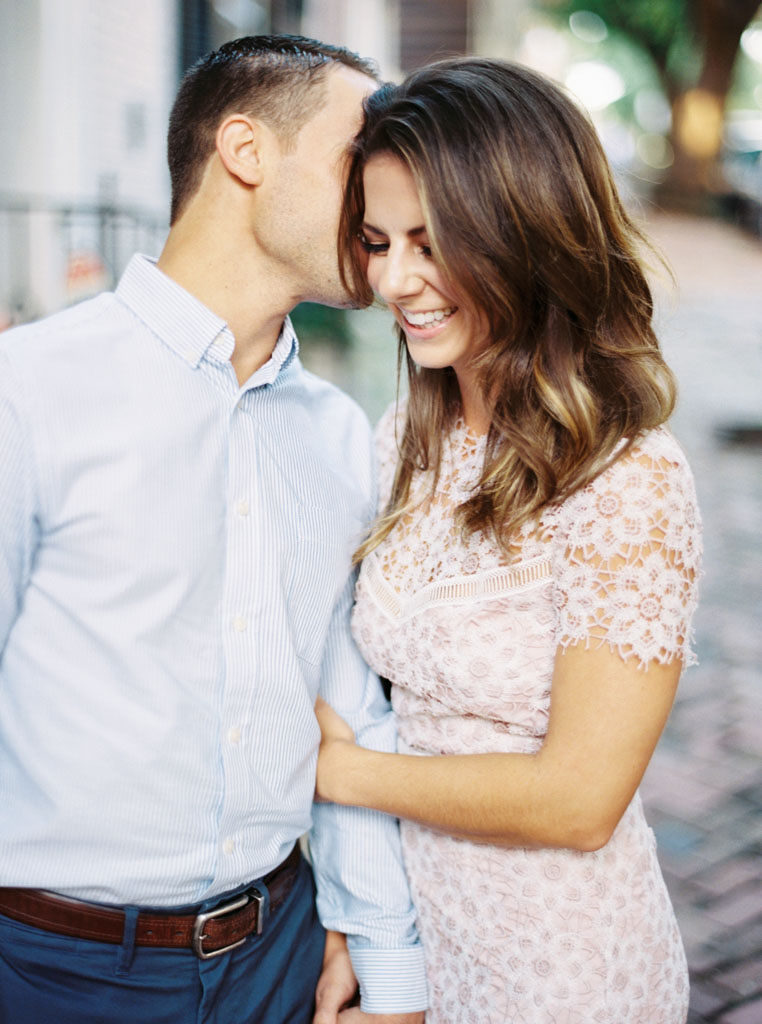 A man kisses his fiance's cheek as she smiles with her eyes closed and holds his arm on a brick pathway in Old Town Alexandria, Virginia. Photographed by Virginia wedding photographer Kim Branagan.