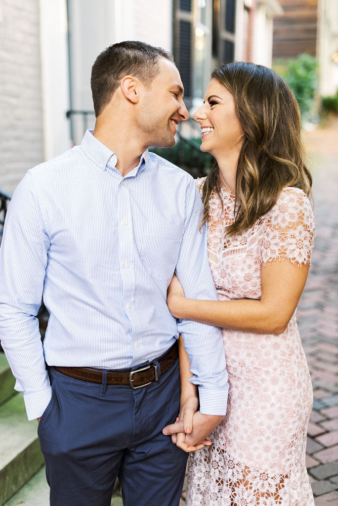 An engaged couple holds hands and gazes into each other's eyes, laughing, on a brick pathway in Old Town Alexandria, Virginia.