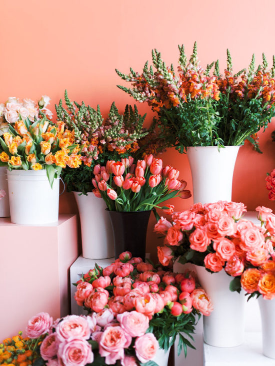 Large bunches of flowers, such as peonies, roses, and tulips, in white vases on the ground in front of a coral-colored wall. The flowers are all different colors, including different shades of pink, yellow, and orange. Photographed by Virginia commercial photographer Kim Branagan at Sweet Root Village in Alexandria, VA.