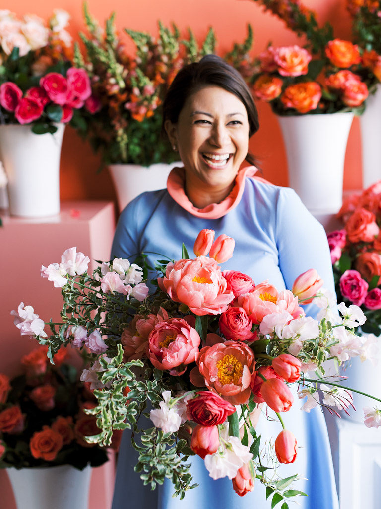 A woman wearing a blue dress smiles and looks off into the distance as she holds a lush bouquet of large, pink peonies, pink tulips, and pops of small, lavender-colored flowers. She is standing in front of a coral pink wall and several large vases full of pink and red flowers.