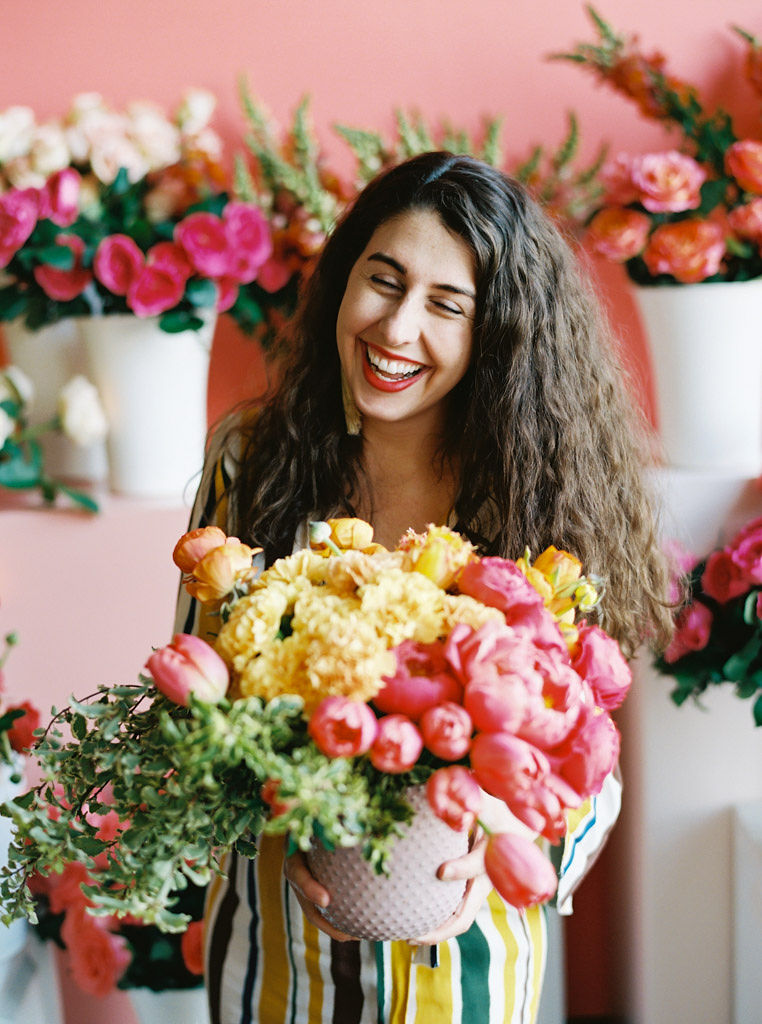 A woman with long and curly brown hair laughs as she holds her pastel pink vase full of bright yellow and pink florals, with greens cascading down the side of the arrangement.