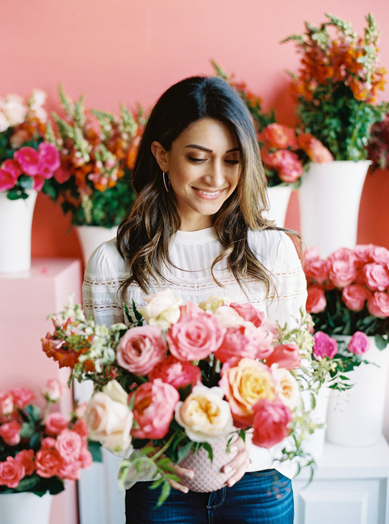 A woman with long, ombre brown hair wearing big hoop earrings a white blouse smiles and looks down at her bouquet of pink and cream peonies with greenery throughout. She is standing in front of a pink wall and big floral bouquets. Taken by Maryland commercial photographer Kim Branagan.