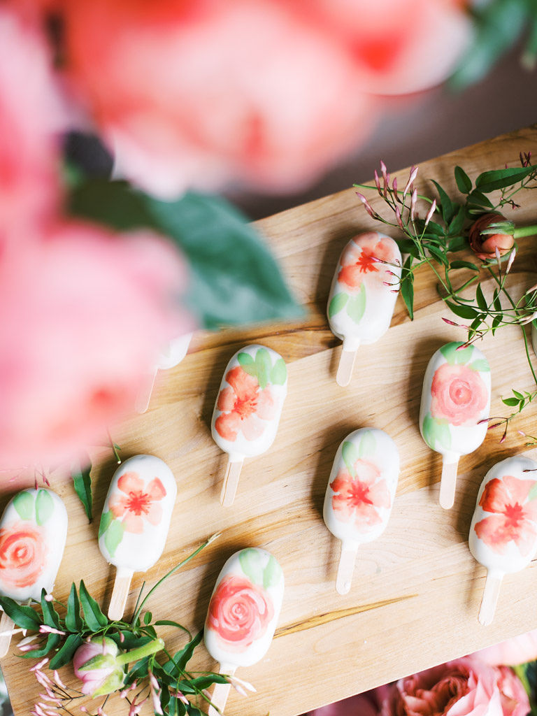 White popsicles with a watercolor like design of a pink flower with green leaves arranged on a wooden board. Loose flowers, stems, and greenery surround the popsicles.