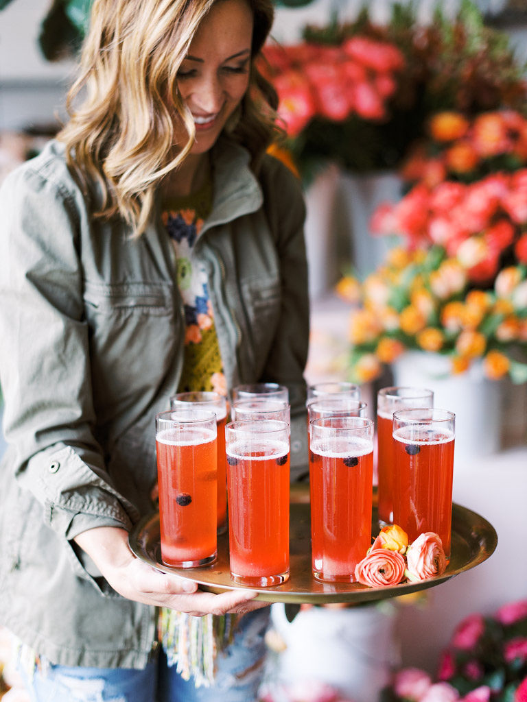 A woman with blonde hair wearing a gray canvas jacket holds a tray of tall glasses filled with a dark pink beverage and fresh blueberries to garnish. Several rose buds lay on the platter. There are lots of pink floral bouquets in the background. Taken at Sweet Root Village in Alexandria, VA.