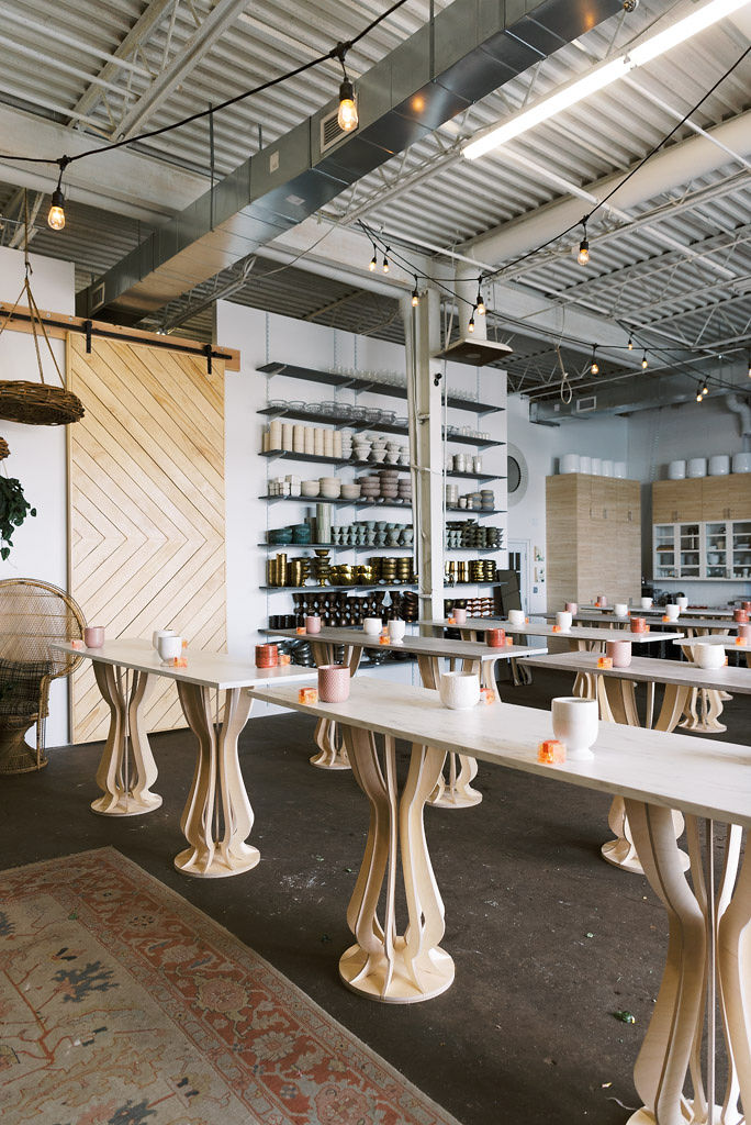 Rows of long, tall wooden tables (bar style) with white and pink vases on them. Christmas bulb lights hang on the ceiling above the tables. A large wooden sliding door and shelves of vases is in the background. Image is of Sweet Root Village, a floral design studio in Alexandria, VA. Taken by Virginia commercial and wedding photographer Kim Branagan.