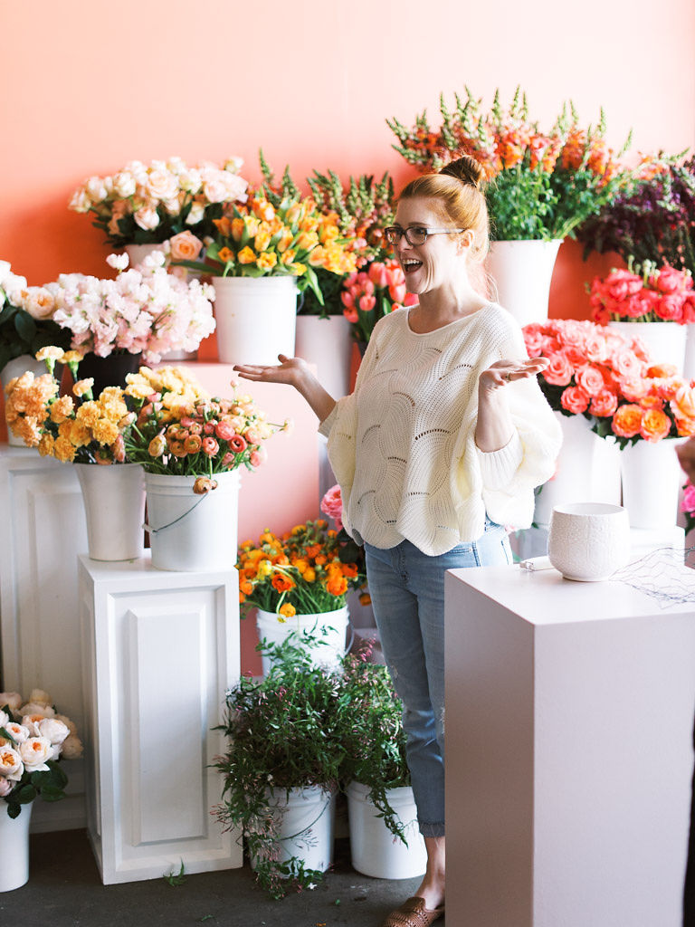An employee of Sweet Root Village teaches attendees of a floral workshop. Large bunches of pink, red, orange, and yellow flowers are behind her. Taken by D.C. commercial photographer Kim Branagan.