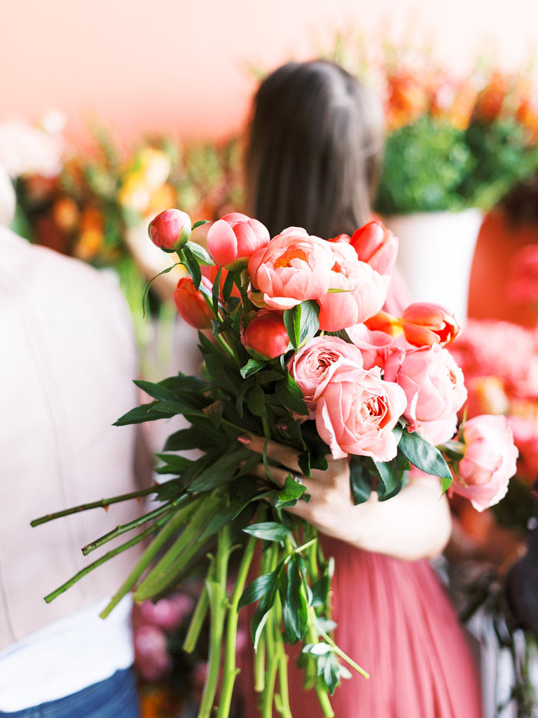Close up shot of a woman's hand holding a bouquet of light pink roses, peonies, and tulips with dark green leaves. The background of the image, where people are walking around many other large bouquets of colorful flowers, is blurred.